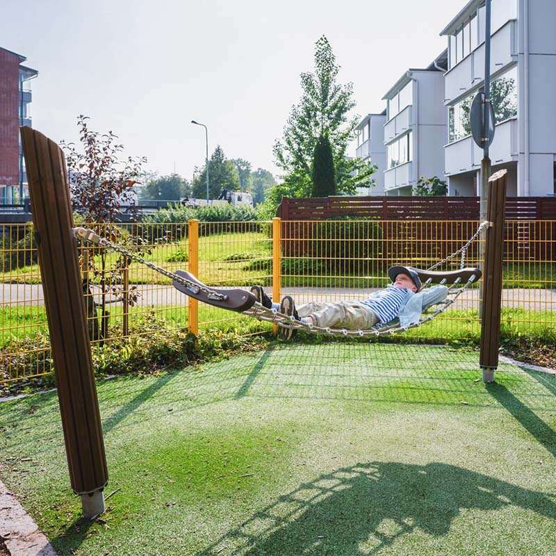 Pheasant themed playground in Tuusula/Boy in a hammock A young boy in a striped shirt and hat lies on a rope hammock-style swing in a playground. The swing is suspended between two wooden posts on a green turf surface. Modern residential buildings and a street with a car are in the background.