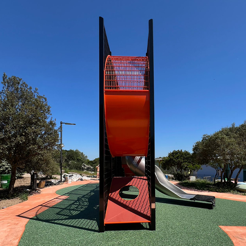 Wall-Holla Park Sardinia 4 A modern playground structure with a tall, rectangular design featuring orange climbing elements and a metal slide against a backdrop of clear blue sky and surrounding trees. The ground is covered with green rubber safety turf and a pathway.