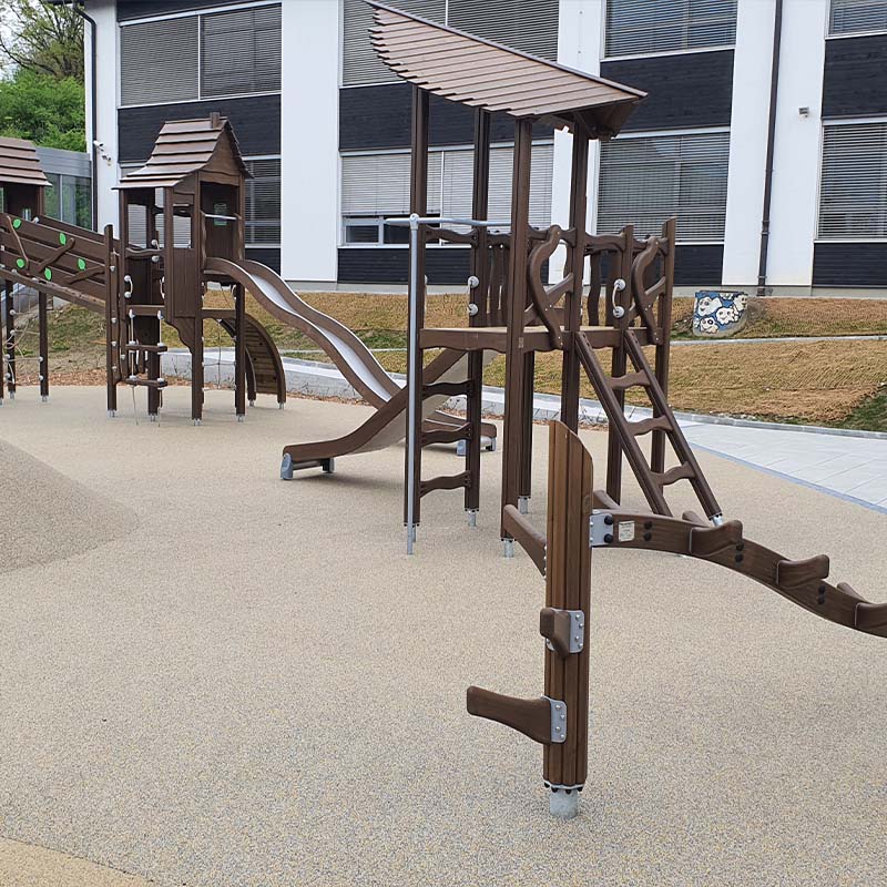 Playground of Škofljica Primary School A modern playground with wooden climbing structures, slides, and ladders on a soft, padded surface. The playground is situated in an open area near a building with large windows. The equipment includes multiple levels and activities for children to enjoy.