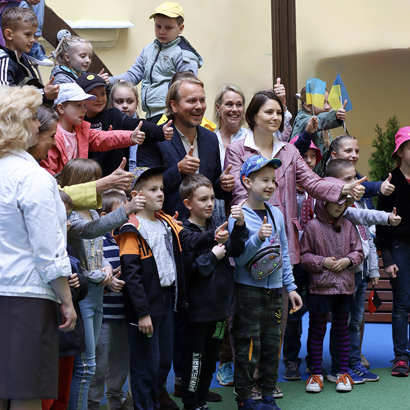 Joy of play Lviv opening group picture 800x800 A group of adults and children stand together in a cheerful setting. They are giving thumbs up and smiling. Many children wear brightly colored clothing and hats. Some hold small flags. The image captures a positive and joyful moment of a community gathering.