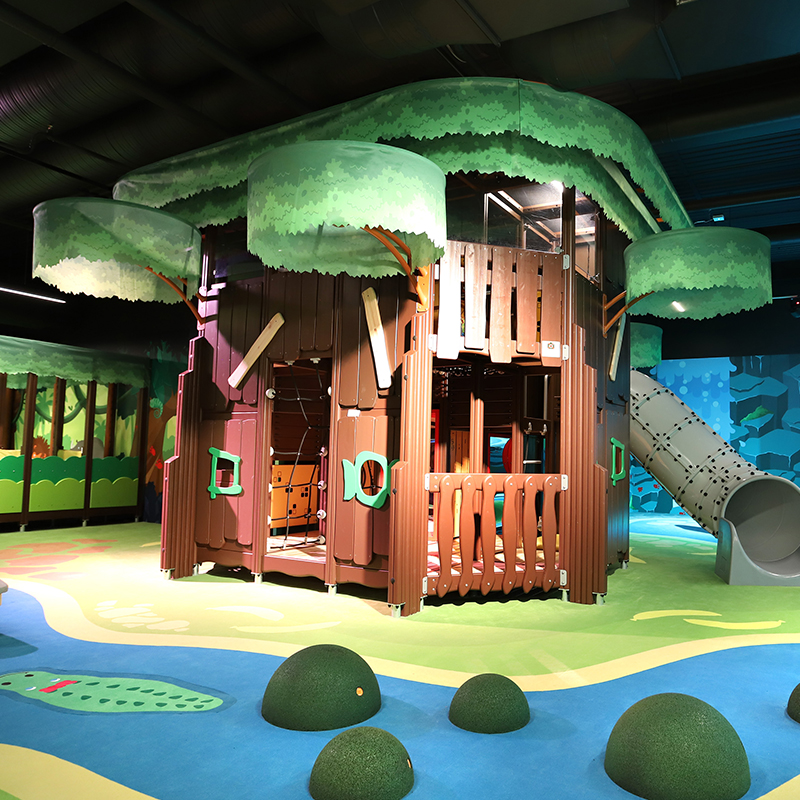 Balubaland 2 Indoor playground featuring a large wooden treehouse with green canopies, various play elements, and slides. The vibrant floor is decorated with colorful patterns and shapes to mimic nature, adding an engaging and playful atmosphere.