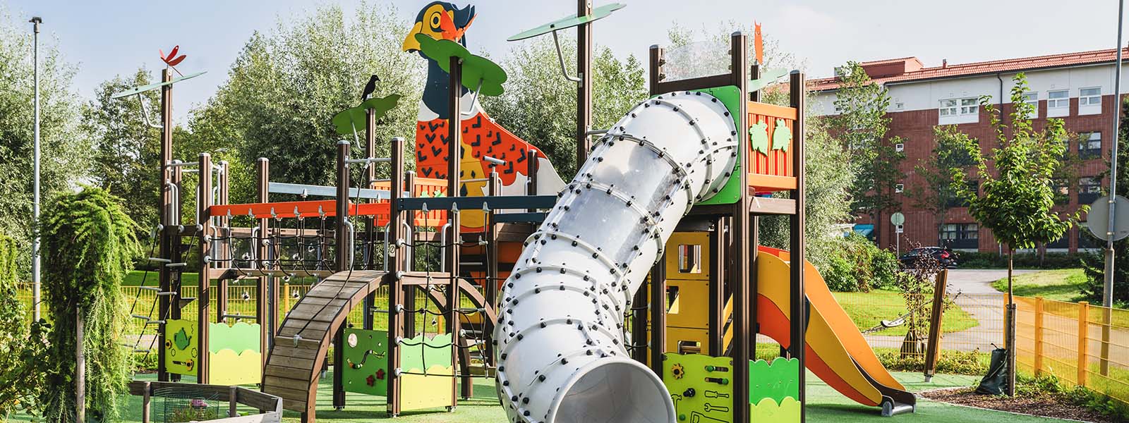 Pheasant themed playground in Tuusula A colorful playground with a large jungle gym featuring a bird design. The structure includes slides, climbing areas, and bridges. Surrounding the playground is green grass, trees, and a few apartment buildings in the background.