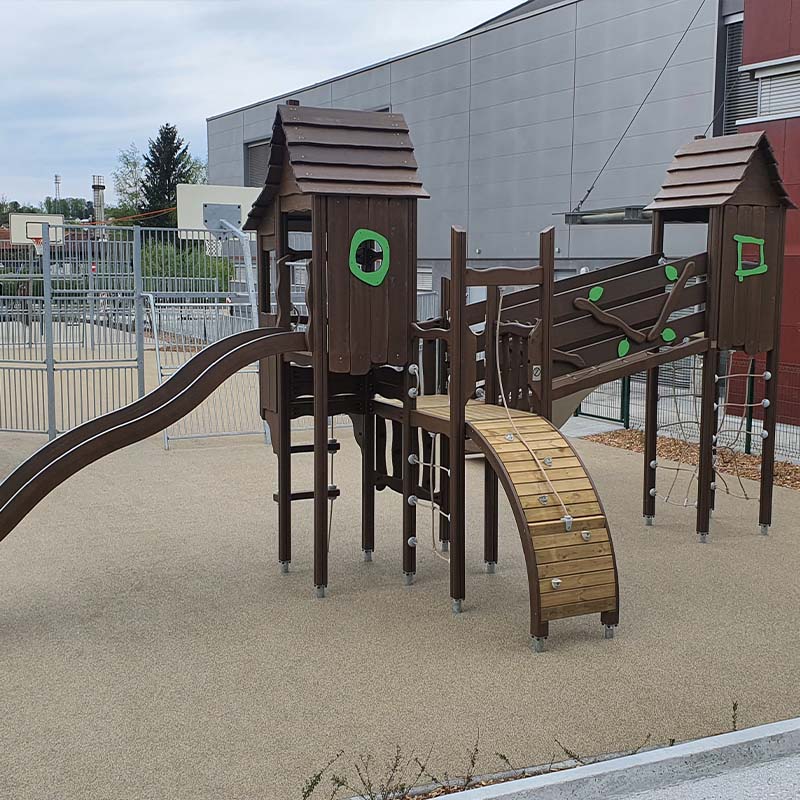 Playground of Škofljica Primary School A modern playground structure featuring two brown playhouses with green details connected by a bridge, slide, and climbing elements. They stand on a soft play surface within a fenced area, with a large building in the background.