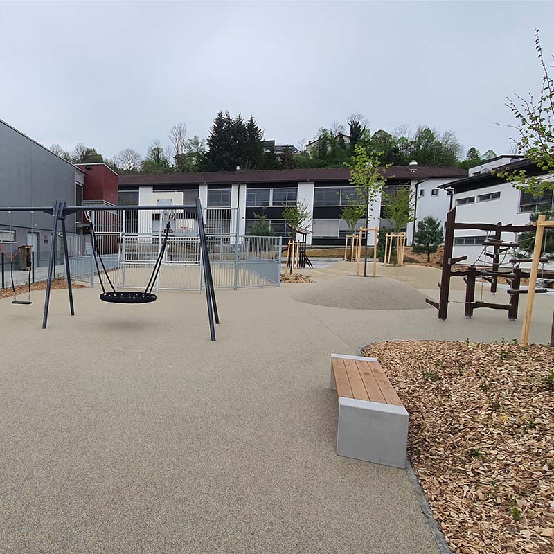 Playground of Škofljica Primary School A modern, outdoor playground featuring a swing set with a basket swing, various climbing structures, and benches. The ground is covered with a smooth, gray surface and a mulch area. The playground is surrounded by buildings and trees in the background.
