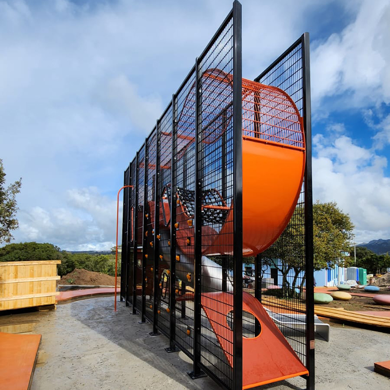 Wall-Holla Park Sardinia 3 A colorful, tall playground structure featuring multiple climbing elements, orange slides, and an enclosed net tunnel. The structure is surrounded by a high black metal frame. The playground is set outdoors on a sunny day, with trees and sky visible in the background.
