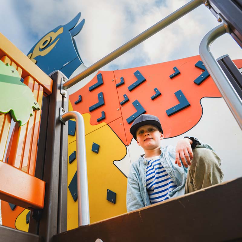 Pheasant themed playground in Tuusula/Boy in the picture A child wearing a striped shirt, denim jacket, and cap crouches at the top of a colorful playground structure with animal shapes and geometric designs. The child looks directly at the camera, and the sky with some clouds is visible in the background.