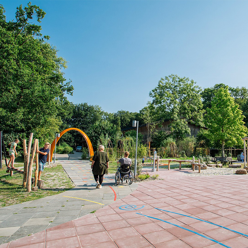 1474 NL Sona Ronald McDonald Hoeve 2022-750800x800 An inclusive outdoor park with a mix of paved pathways, grassy areas, and natural wooden play structures. A person in a wheelchair and another individual walk near an orange archway, while a child plays on a climbing structure. Trees surround the park.
