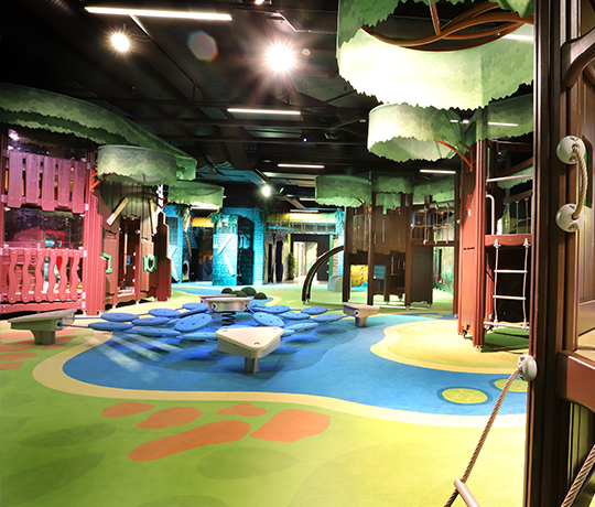 Balubaland An indoor playground with a nature theme featuring artificial trees, wooden play structures, and a blue, river-like flooring. The space has various play equipment, including climbing areas, platforms, and a slide. The lighting is bright, enhancing the vibrant colors.