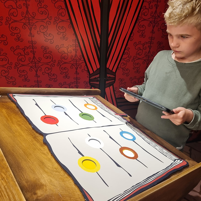 Rio och Aaron LasseMaja 2 800x800 A young boy holds a tablet while engaging with a colorful interactive exhibit. The exhibit resembles a large open book with six differently colored circles on each page. The background shows red and black curtains creating a decorative setting.