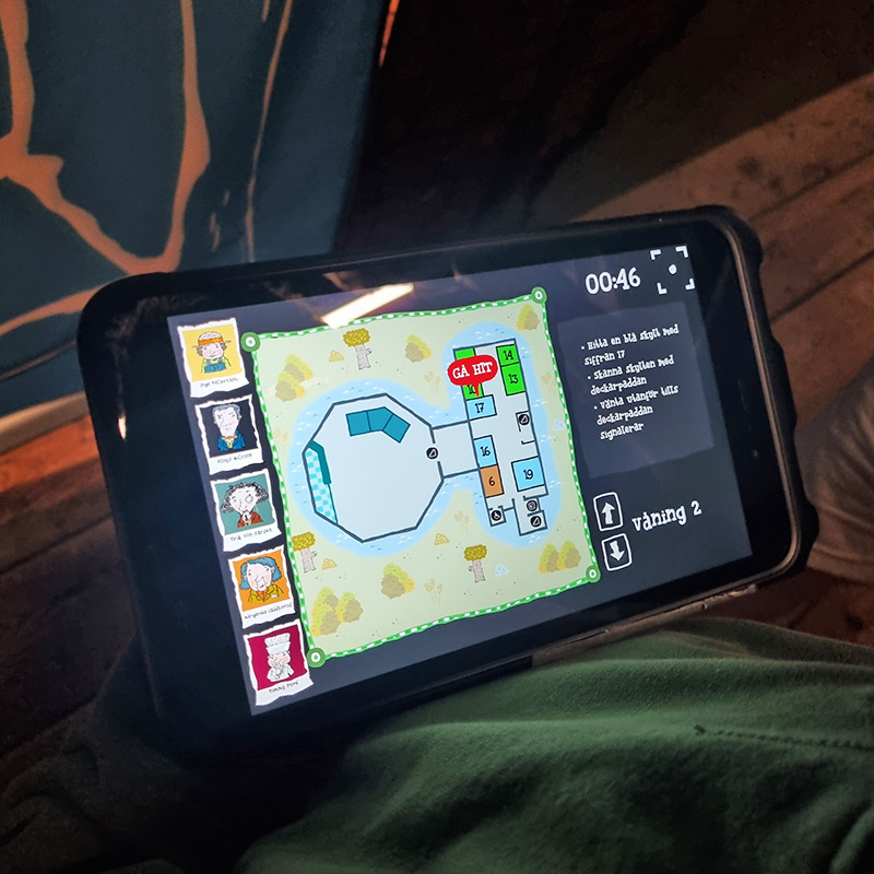 Rio och Aaron LasseMaja 6 800x800 A person is holding a phone displaying a game map interface. The map shows various rooms and characters, with a timer at the top right corner and text, likely instructions, beside it. The environment appears to be dimly lit.