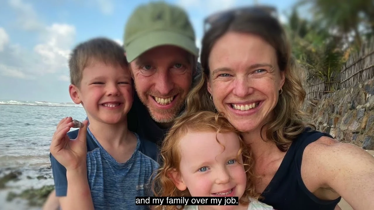 Two adults and two children posing together at a seaside location with ocean waves, a stone wall, and trees in the background.