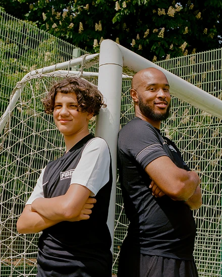 Smiling young boy and man standing back to back with arms crossed in front of a soccer goal