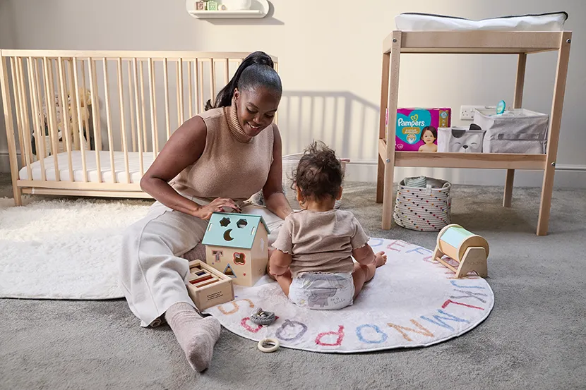 A woman sitting on the floor in a nursery playing with a toddler who is wearing a diaper. They are surrounded by wooden toys, with a crib and a changing table in the background.