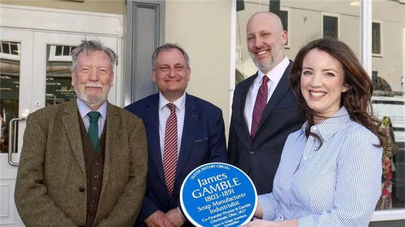 Four people standing outside a building holding a James Gamble heritage plaque marking the co-founder of Procter & Gamble.