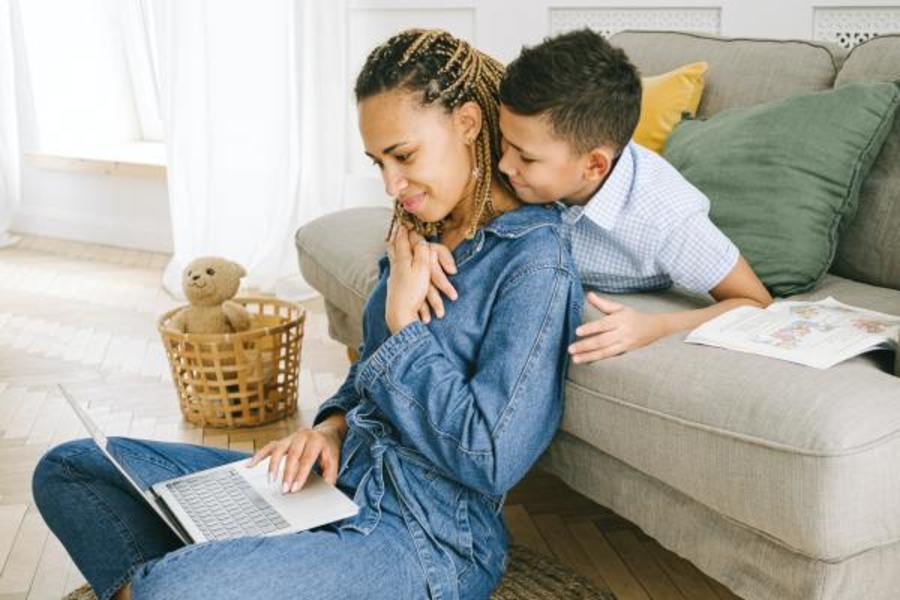 woman sitting on floor in front of couch with little boy looking over her shoulder while she is on her laptop.