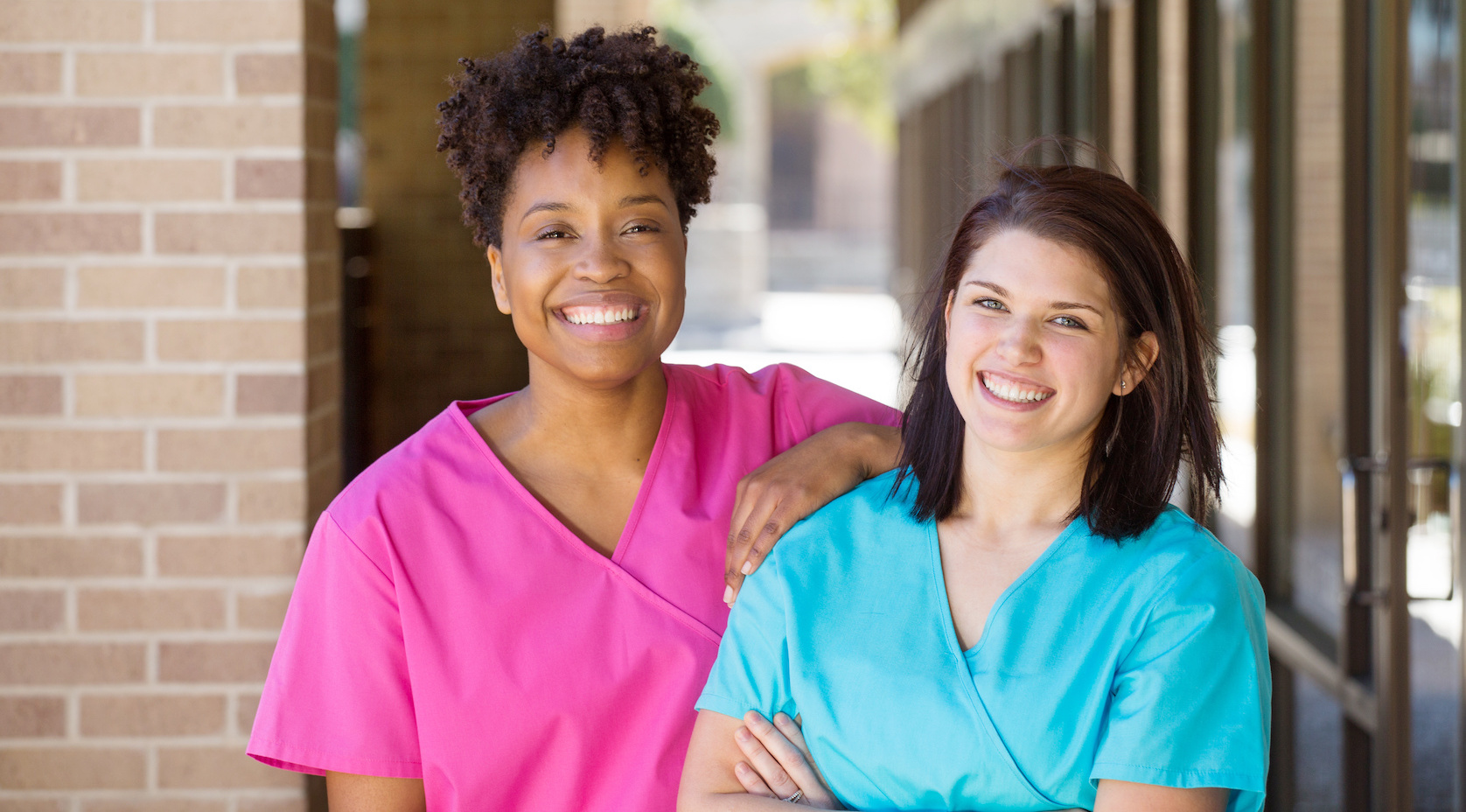 Two smiling Direct Primary Care Providers in pink and turquoise medical scrubs standing outside a DPC practice.