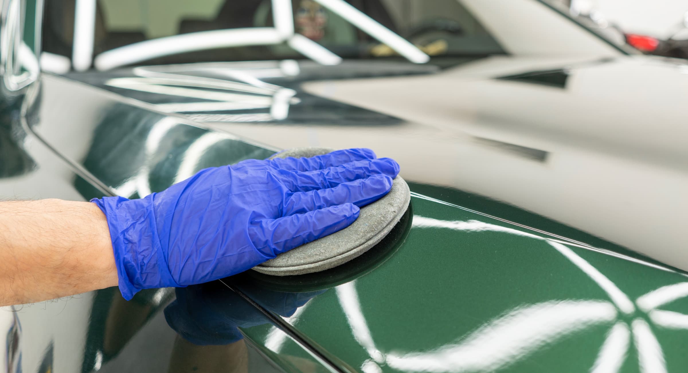 Close-up of a gloved hand rubbing a puffing pad on the hood of a shiny, green car.