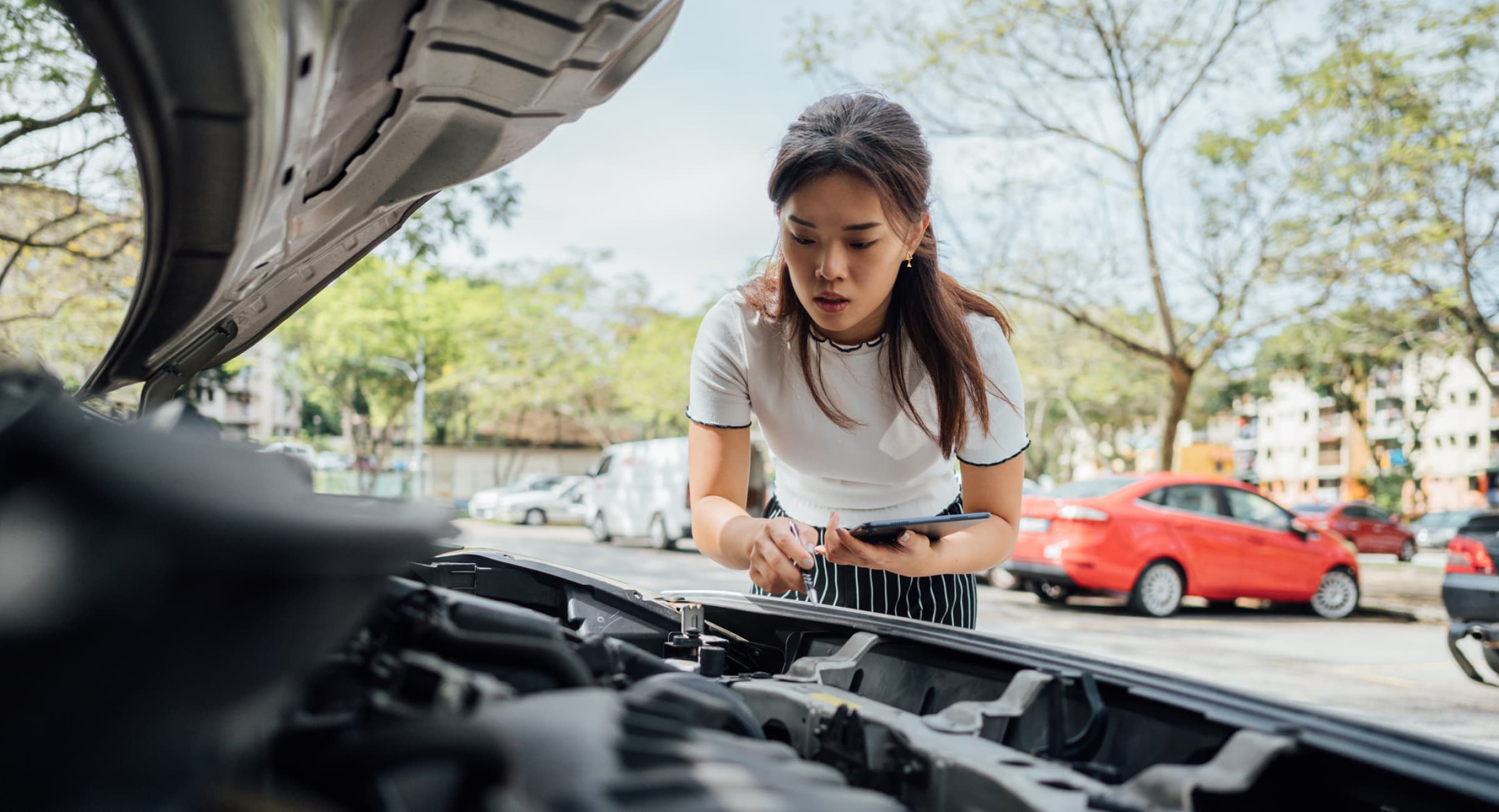 A woman holding a notepad and pencil looks under the hood at a car's engine. 