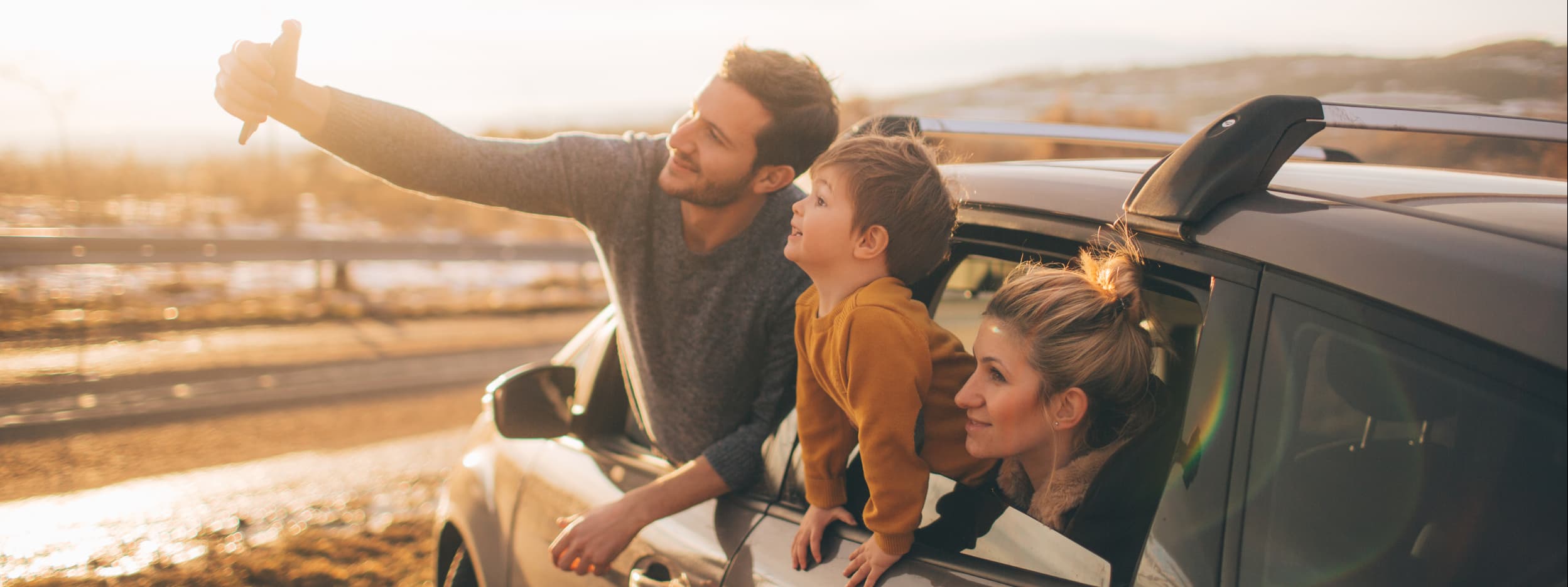 A smiling man takes a selfie of himself, a young child, and a woman as they lean out of the windows of a car parked on a beach at sunset.