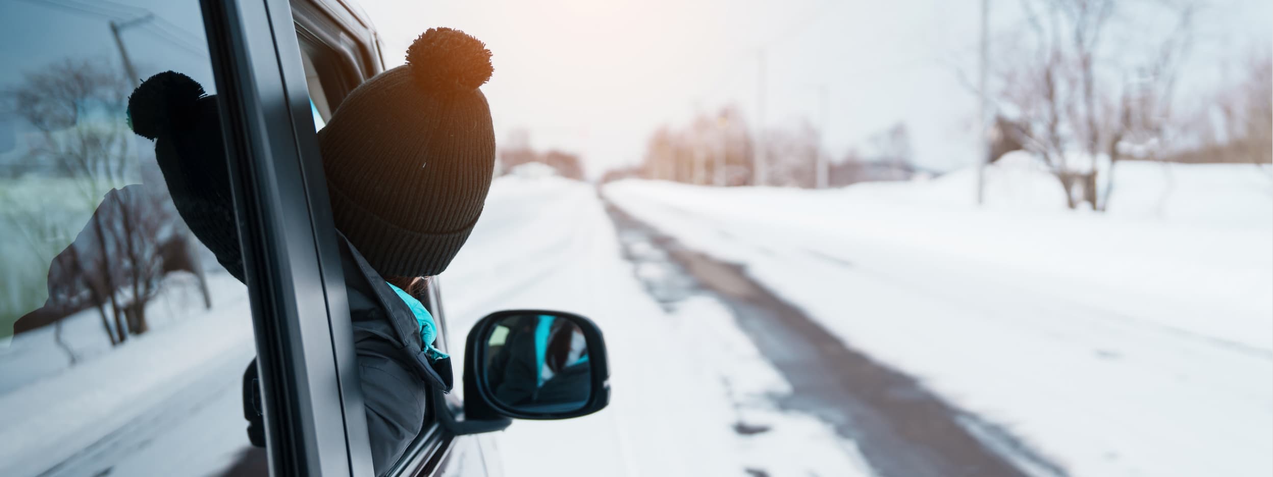 A person in a winter coat and hat leans out of the passenger window of a car to look at the snowy road ahead. 