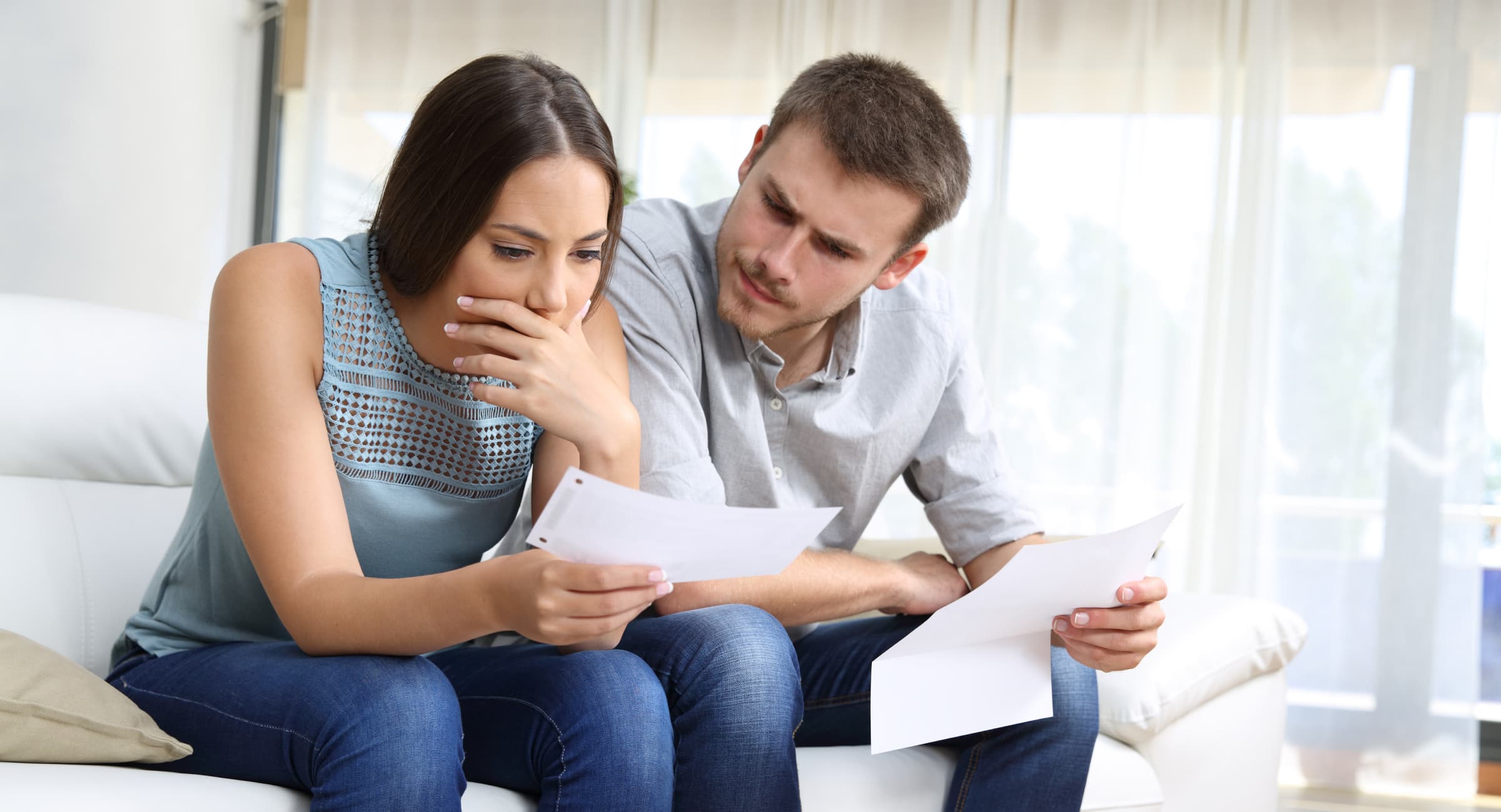 A woman holding an auto insurance bill and the man sitting next to her on the sofa look concerned.