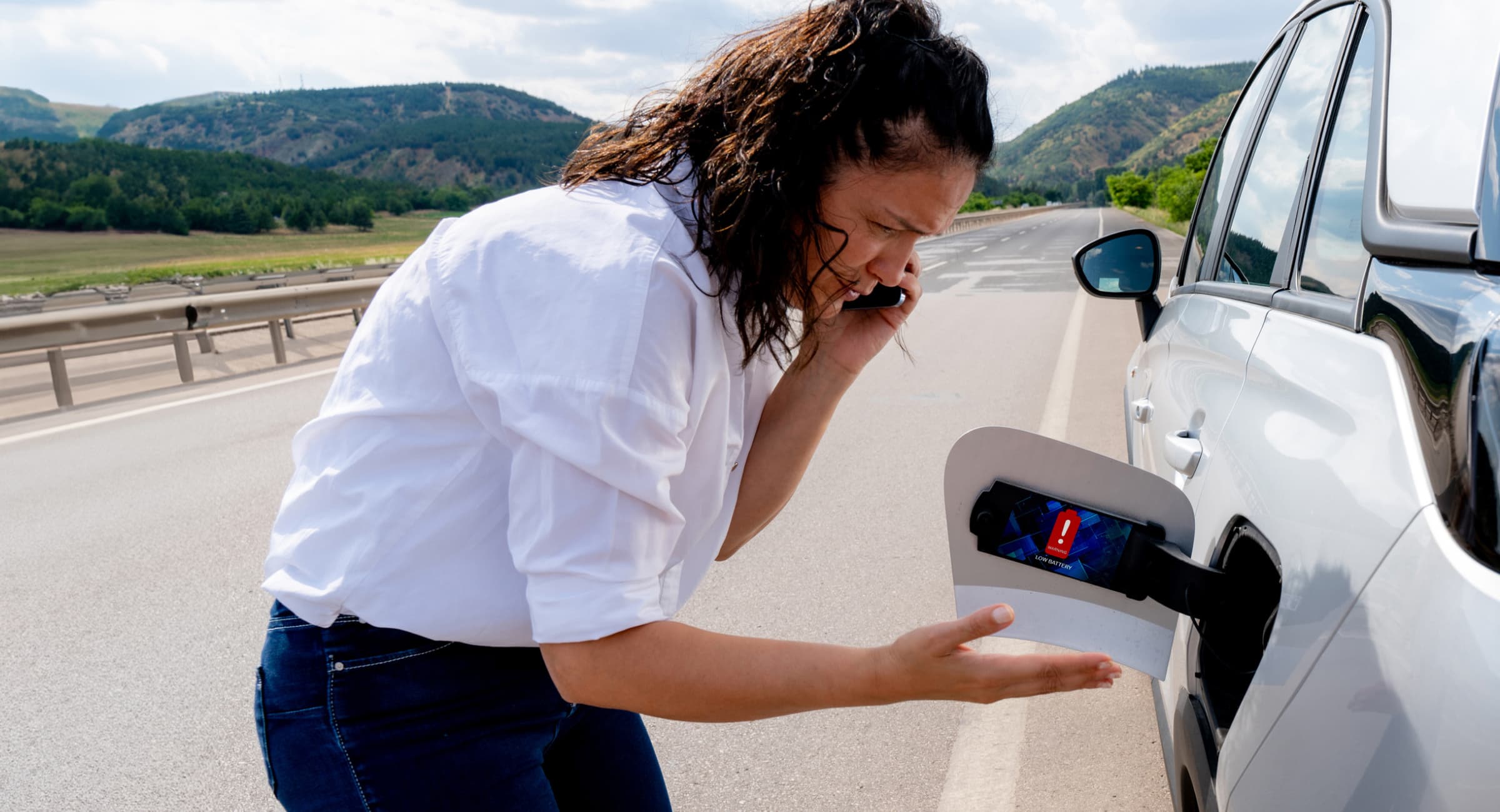 A frustrated-looking woman speaking on her mobile phone looks at the charging port of her electric car which is pulled over on the side of the road.