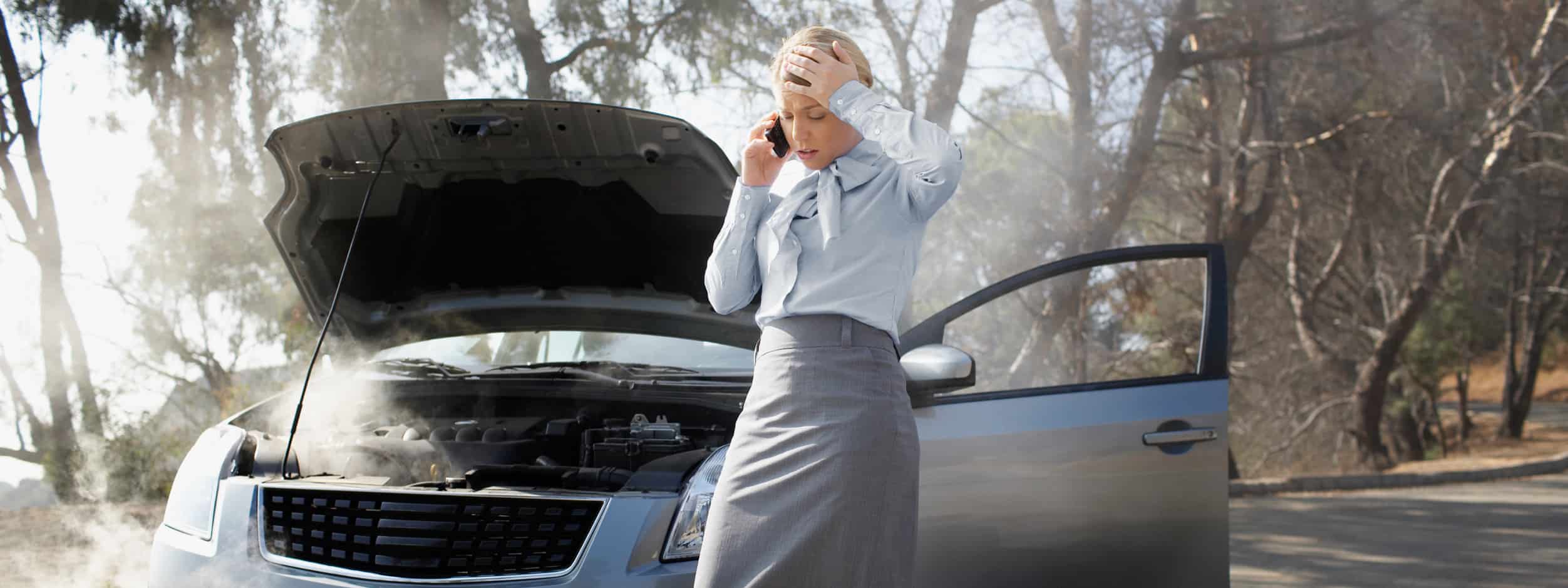 A woman stands in front of her overheating car while on the phone.