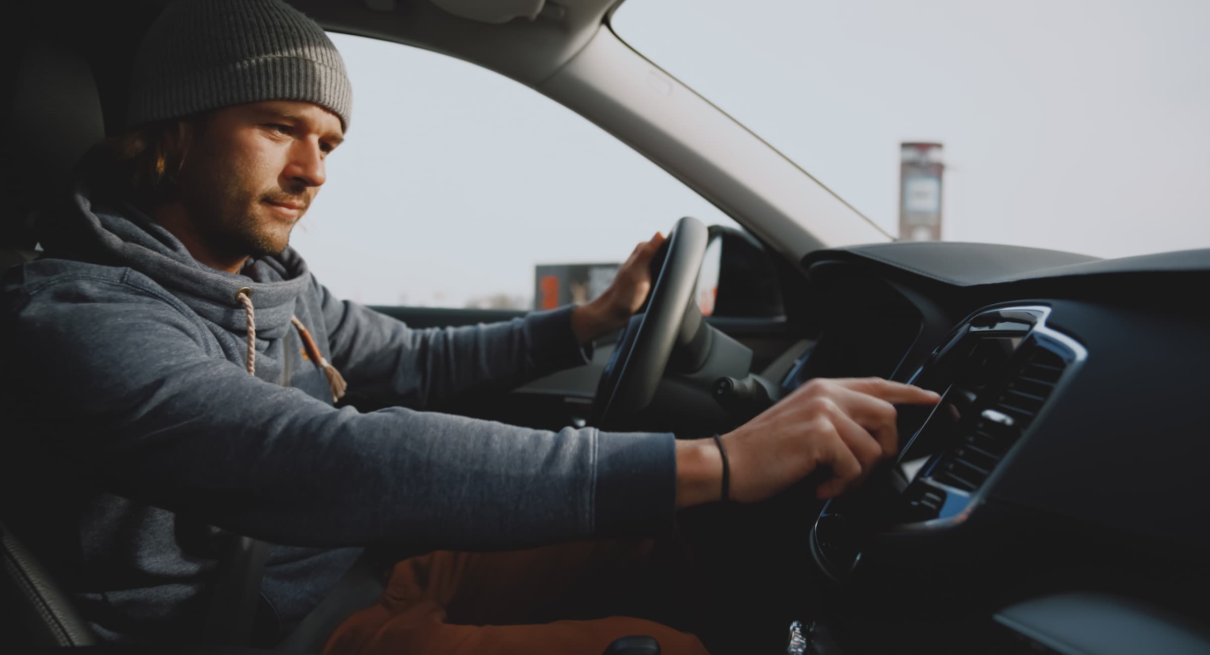 A man sitting in his car is reaching for touch screen on the vehicle's infotainment system.