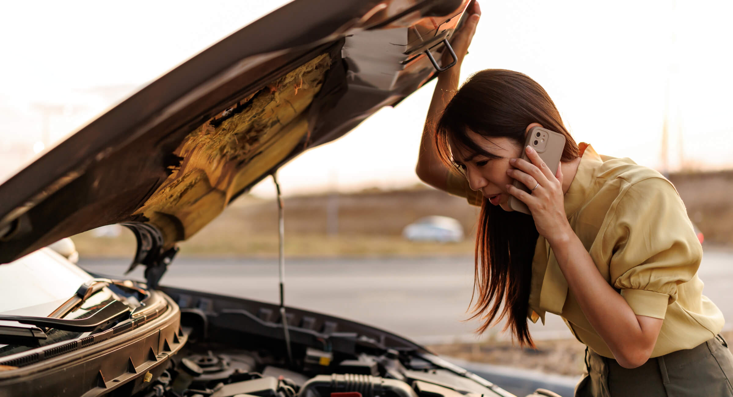 A woman talks on the phone while looking into the engine compartment of her car, which is stopped on the side of the road. 