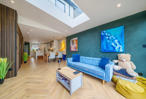 A large, contemporary and colourful open-plan room in an eco home in London. A blue sofa sits against a turquoise wall and herringbone flooring leads towards a modern breakfast bar and kitchen with skylights above.