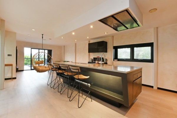 A large and contemporary open plan kitchen, architecturally designed with wooden flooring and high-end fixtures and fittings. A kitchen island sits in the middle of the room with bar stools, in front of a hanging egg chair and doors to the terrace.
