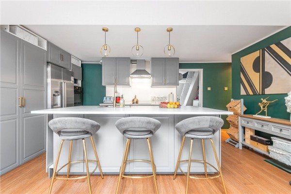 The open plan kitchen of the 10th most viewed property on Zoopla in 2023. There is a white kitchen island with 3 grey and gold bar stools, grey kitchen cabinetry, wooden floors and emerald green walls. 