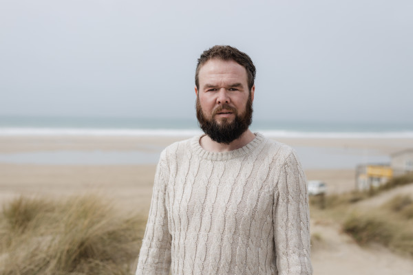 Paul David Smith, a photographer, stands in front of a sandy beach wearing a woolly jumper