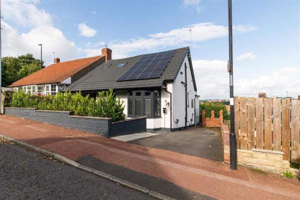 A semi-detached eco bungalow with solar panels on the roof. A tarmac driveway sits to the right of the white house, with a small walled garden in front of the home. Views stretch into the distance behind the house.