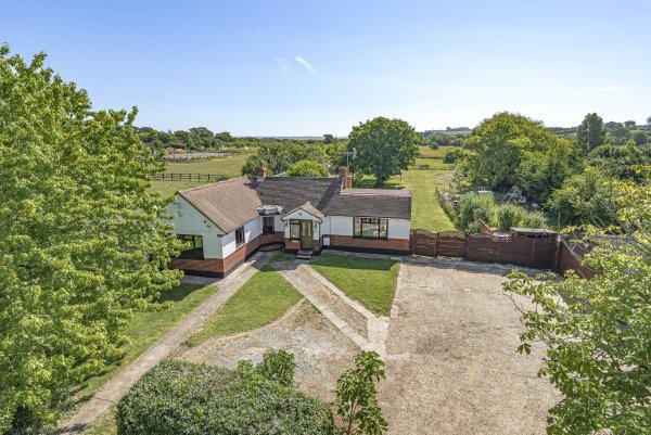 Aerial view of a four bedroom equestrian property in Essex. A large bungalow is fronted by a large gravel driveway and surrounded by mature trees. Past the house you can see lawned gardens and a paddock in the distance.
