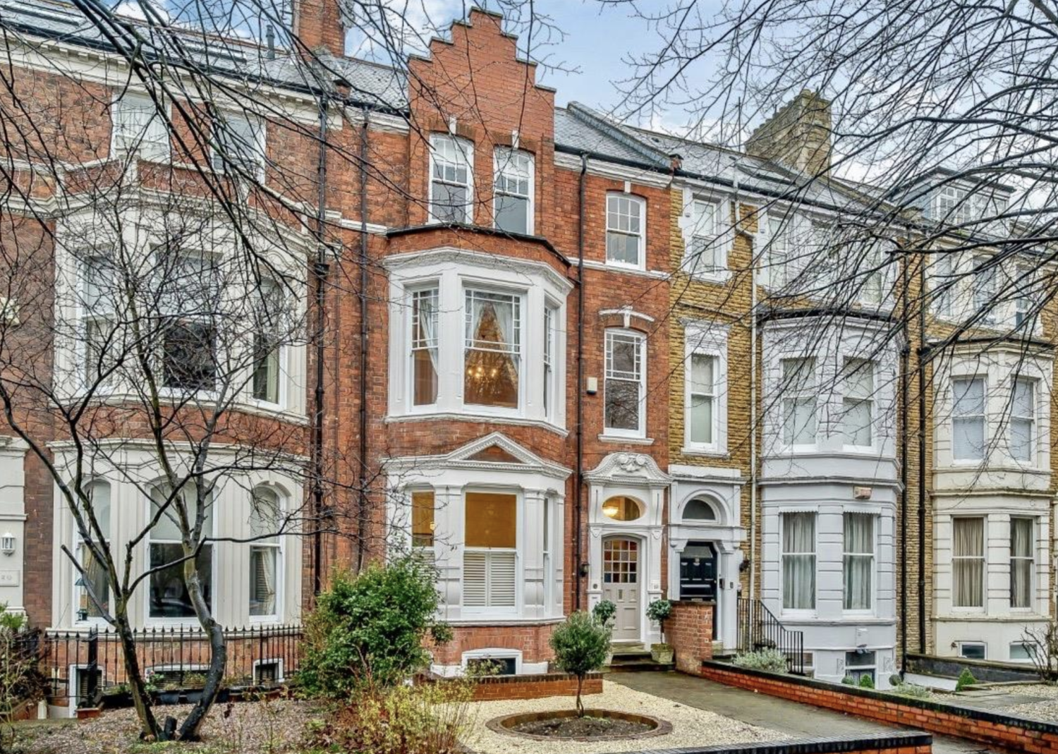 Large red brick Victorian townhouse with bay windows and a front garden with small shrubs and paved area