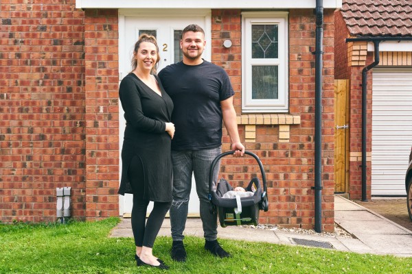 A couple standing outside a red brick house with their newborn baby