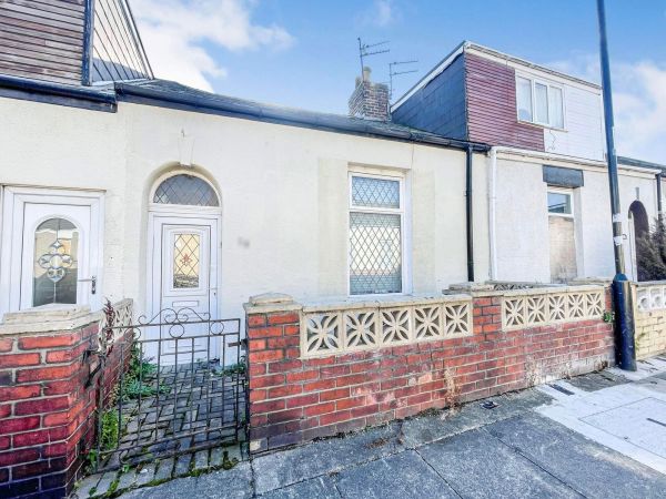The exterior of a small one bedroom cottage for sale in Sunderland. Its small front garden is bordered by a low red-brick wall and a brick garden path leads to the white PVC front door. The one-storey terraced home is white with one window at the front.