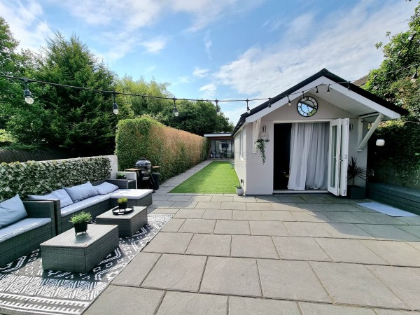 A detached and modern annexe sitting halfway down the garden of a home in Cardiff. There is an outdoor lounge area to the left of the patio and the annexe has double doors with white long curtains wafting out. At the end of the lawn is another outbuilding.