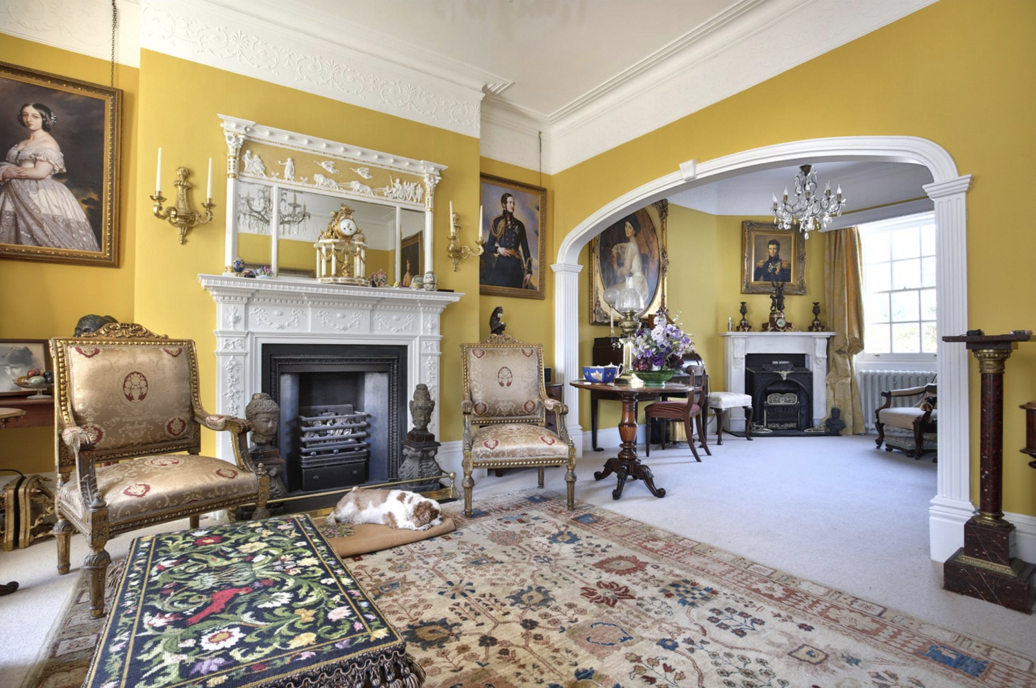 Interior of a Regency-era drawing room painted in mustard yellow, with cornicing and gild-framed paintings and chandeliers hanging above
