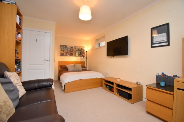 A rectangular bedroom annexe with cream carpet and walls and wooden furniture. There's a double bed in the corner, a TV mounted on the wall and a brown leather sofa facing it.