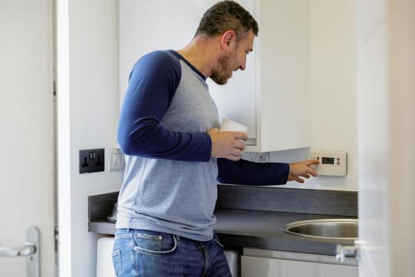 A man holding a cup of tea and pressing a button on the thermostat in his home