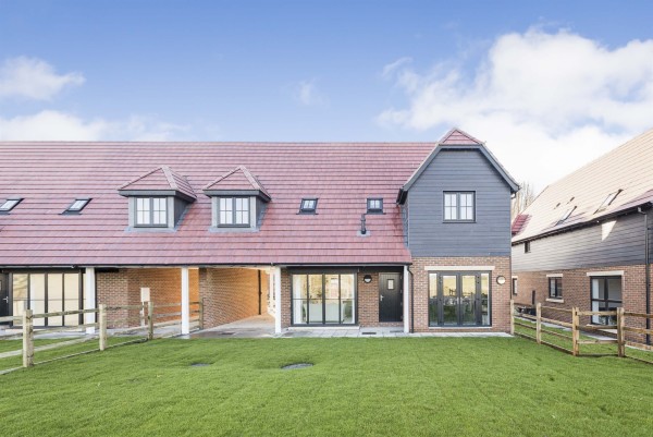 A large lawned garden in front of a terraced new-build home in a barn conversion style, with a covered driveway, french doors, wooden wall feature and red tiled roof