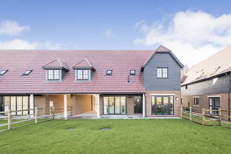 A large lawned garden in front of a terraced new-build home in a barn conversion style, with a covered driveway, french doors, wooden wall feature and red tiled roof