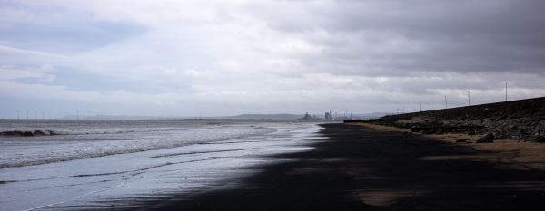 Seaton Carew beach near Hartlepool below a moody sky. The sea on the left looks grey and cold with small waves creeping up the dark sand beach. You can see wind turbines and an industrial port in the distance.