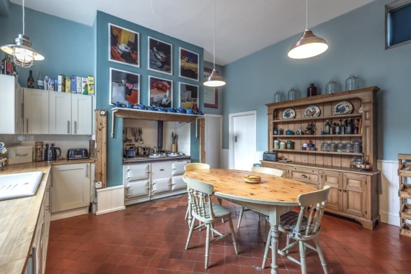 A large country kitchen in a 13 bedroom house in Powys, Wales. The kitchen has a cream Aga, a large wooden dresser, a scrubbed wooden table in the centre with chairs and a terracotta tiled floor.