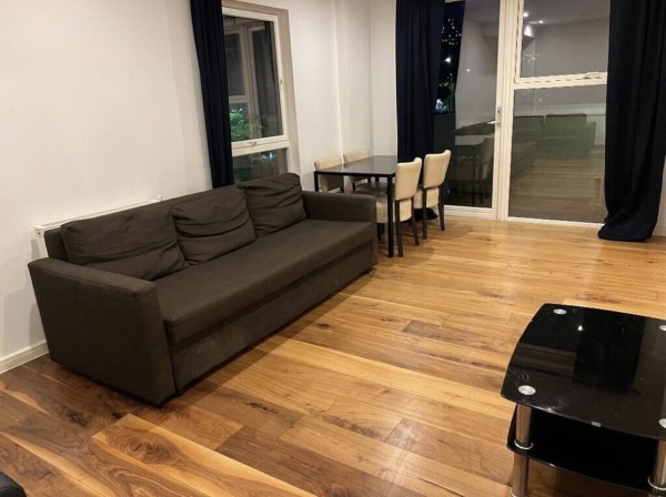 The lounge of a buy-to-let flat in central London. It has a hardwood floor and some basic furniture - a simple brown sofa, a black glass TV stand and a small dining table with chairs.