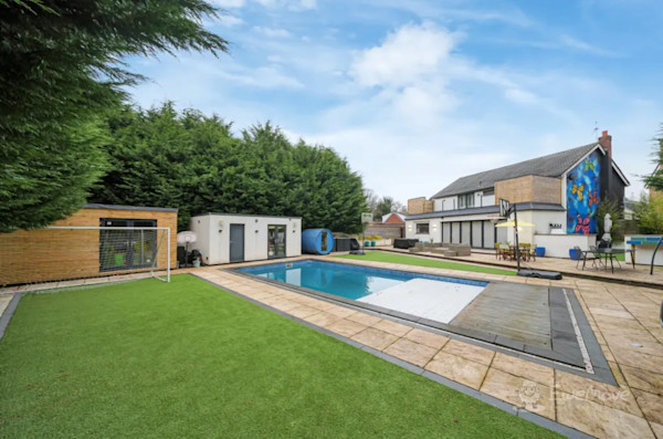 The back garden of a modern house with a swimming pool, some outbuildings and a large patio area
