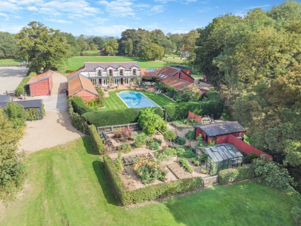 Aerial view of an equestrian property in Suffolk. A large house with several adjoining buildings has a grand swimming pool in the mature landscaped gardens. There is a vegetable patch area with a greenhouse and sprawling lawns beyond.
