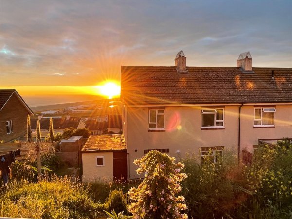 A semi-detached house on a hill with the sun setting in the distance. The view behind the house stretches to the coast. 